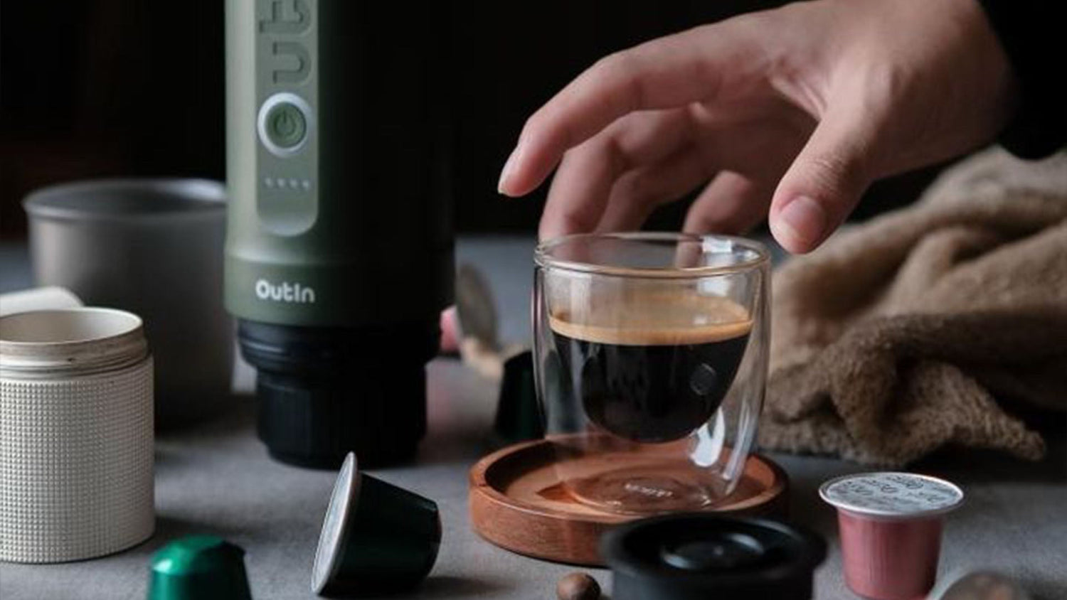 Hand reaching for a double-wall espresso glass next to a portable Outln coffee maker, demonstrating convenient at-home coffee alternatives to instant coffee for on-the-go brewing